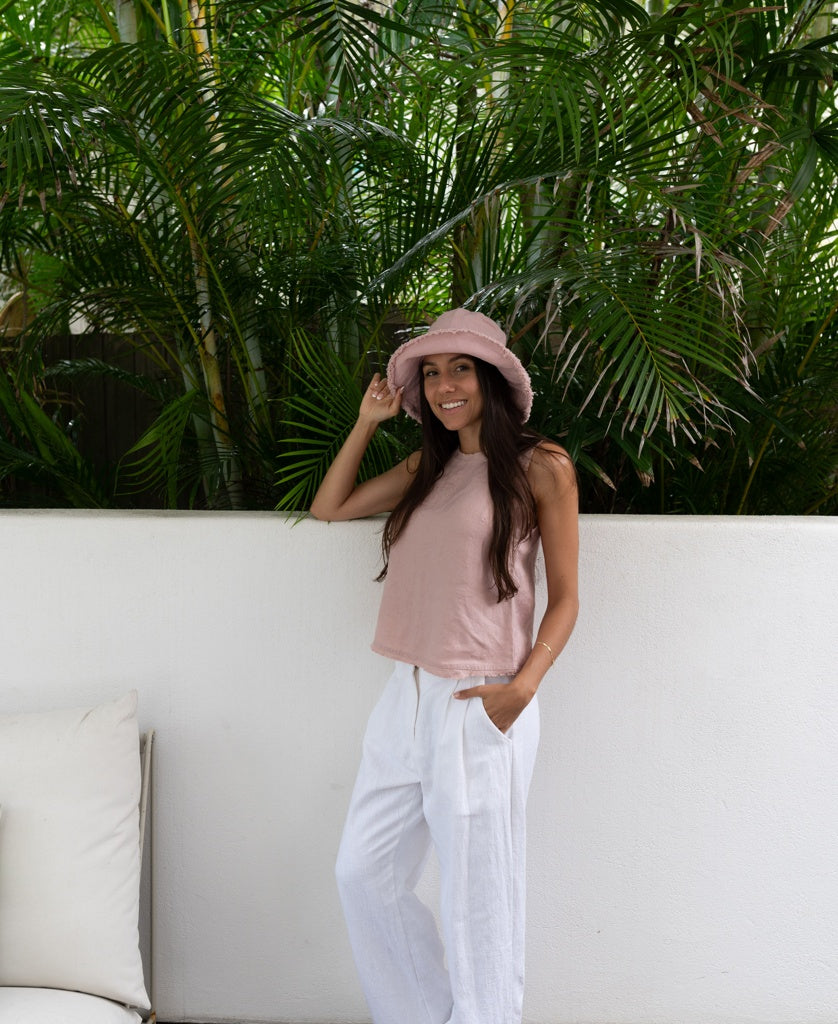 A woman stands by a white wall with green plants, smiling with one hand in her pocket. She wears the Lazy Beach Halo Bucket Hat in Blush, a sleeveless pink top, and white pants.