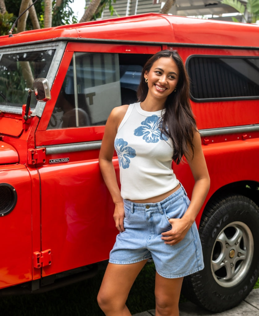A woman smiles beside a vintage red Land Rover, wearing the Rosa Hibiscus Tank in Azure by Golden Breed and denim shorts.