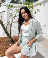 A woman with long dark hair smiles while sitting outdoors on a bench, wearing the Lazy Beach Maeve L/S Shirt in Olive Stripe and shorts over a white tank top.