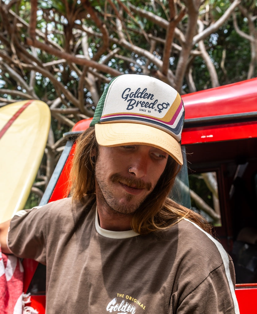 A man with long hair and a mustache wears the Golden Breed Retro Trucker Cap in Wash Orange and a brown tee, standing by a red van and surfboard. Trees are visible in the background.