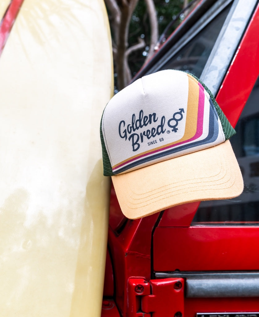 A Golden Breed Retro Trucker Cap in Wash Orange with stripes and an adjustable strap rests on the side mirror of a red vehicle beside a yellow surfboard.