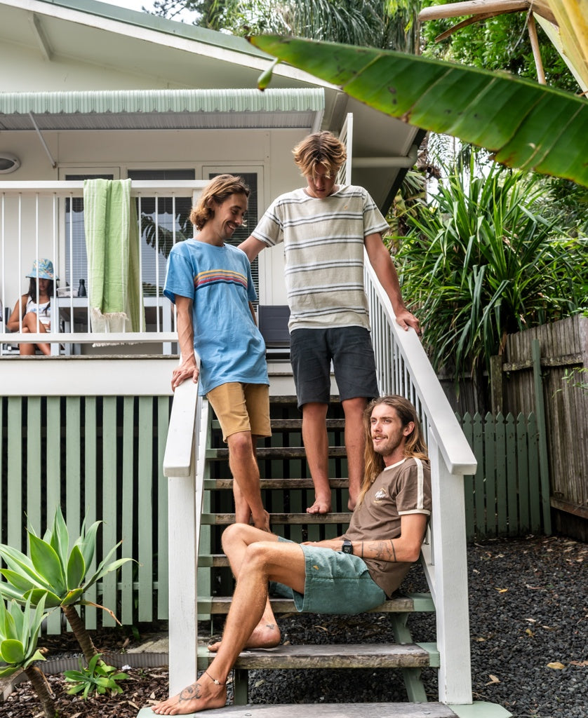 Three people—some in Golden Breed’s Bay Stripe Tee (Mocha) and casual cotton—gather on outdoor steps amid greenery, while a fourth relaxes on the porch, capturing the laid-back Golden Breed spirit.