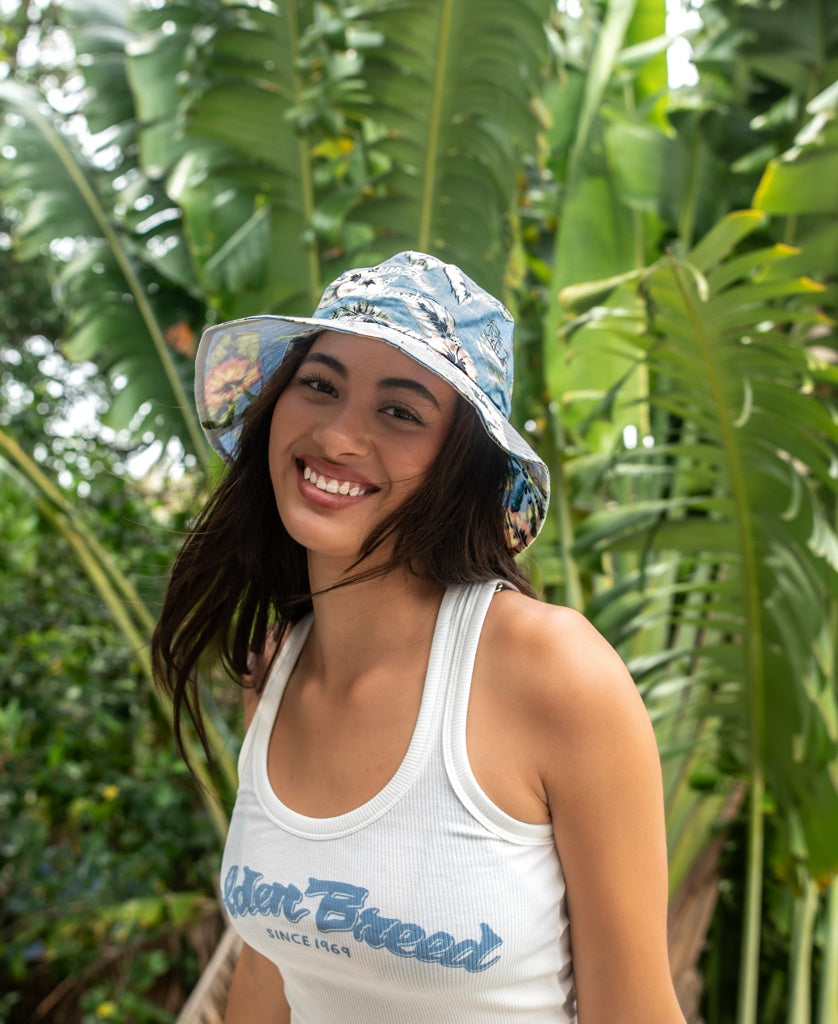 A woman wearing the Golden Breed Chelsea Bucket Hat in Azure Blue and a white tank top smiles in front of large green tropical leaves.