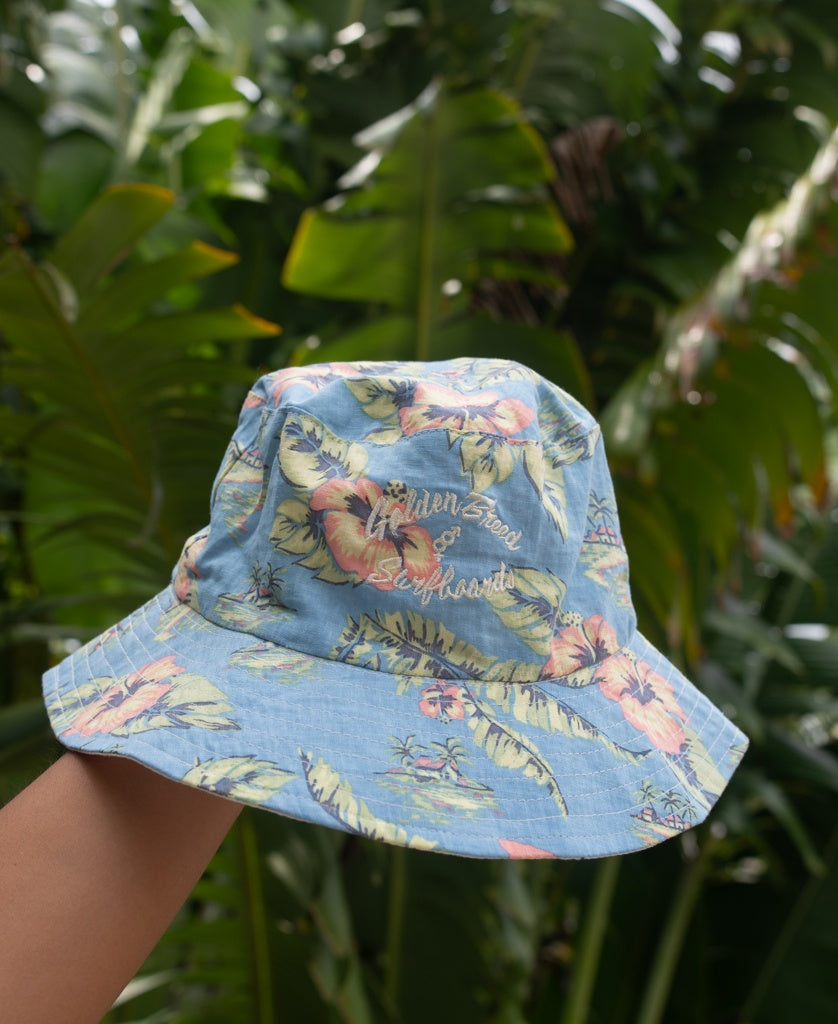 A hand displays the Golden Breed Chelsea Bucket Hat in Azure Blue, featuring a blue floral pattern and embroidered text, against lush green tropical leaves.
