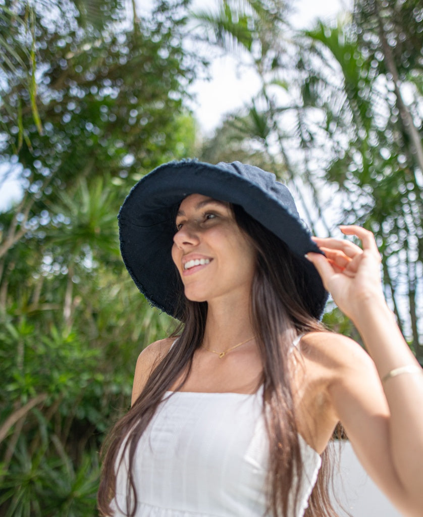 A woman smiles outdoors among green trees and plants, wearing a white cotton dress and the Lazy Beach Halo Bucket Hat in black.