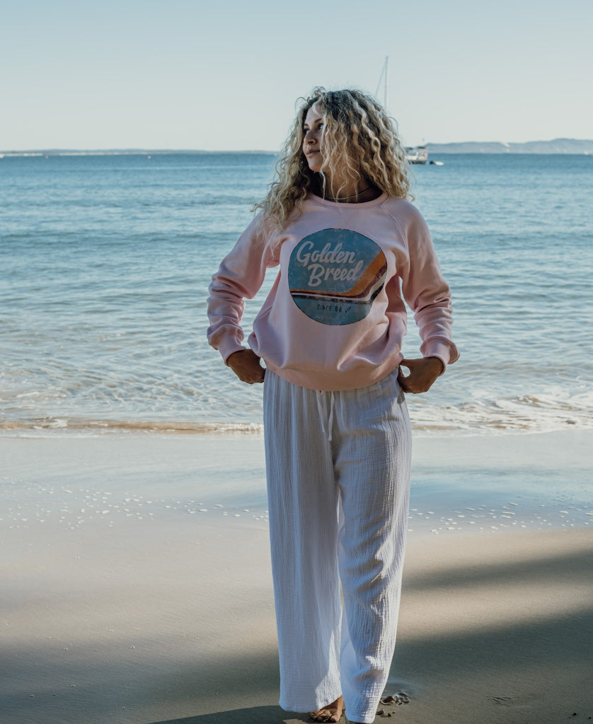 A woman stands on a sandy beach near the water, wearing the Golden Breed Vintage Sweat in Pale Pink, paired with white pants. A sailboat and distant shoreline add to the classic coastal backdrop.
