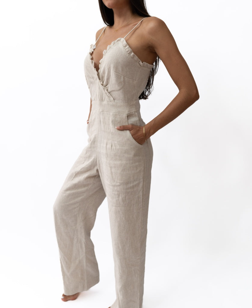 A woman stands with hands in pockets against a white background, wearing the Lazy Beach Emmy Overall | Natural—a handmade in Bali linen jumpsuit featuring thin straps, ruffle details, and wide-leg pants.