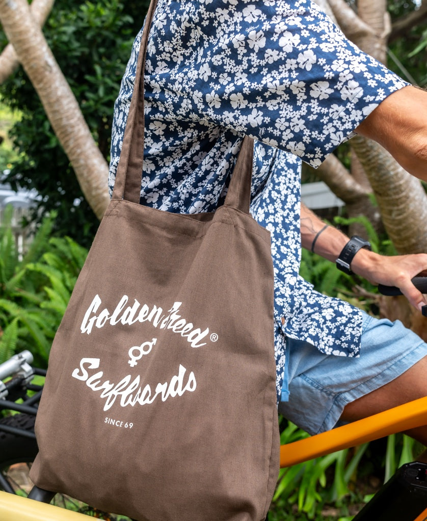 A person in a blue floral shirt and shorts rides a yellow bike, carrying a brown Surf Logo Tote Bag by Golden Breed.