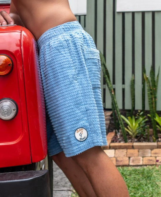 A person wearing Golden Breeds El Solito Walkshort in Azure Blue leans against a red vehicle outdoors, with green plants and a striped fence in the background.