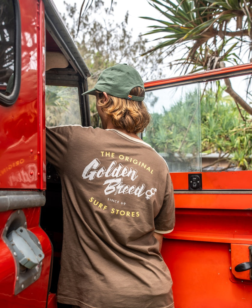 A person in a green cap and Golden Breed Break Ringer Tee in Wash Brown stands by an open red car door, with palm trees in the background.