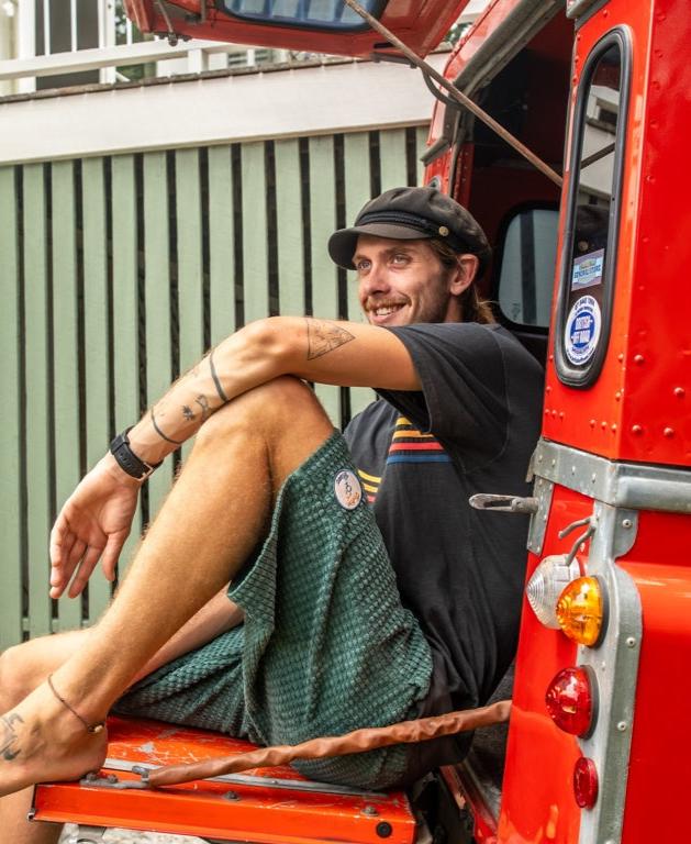 A man in a black cap, T-shirt, and Golden Breed El Solito Walkshort in Ivy Green sits on the open tailgate of a red vehicle, smiling with his arms resting on his knees.