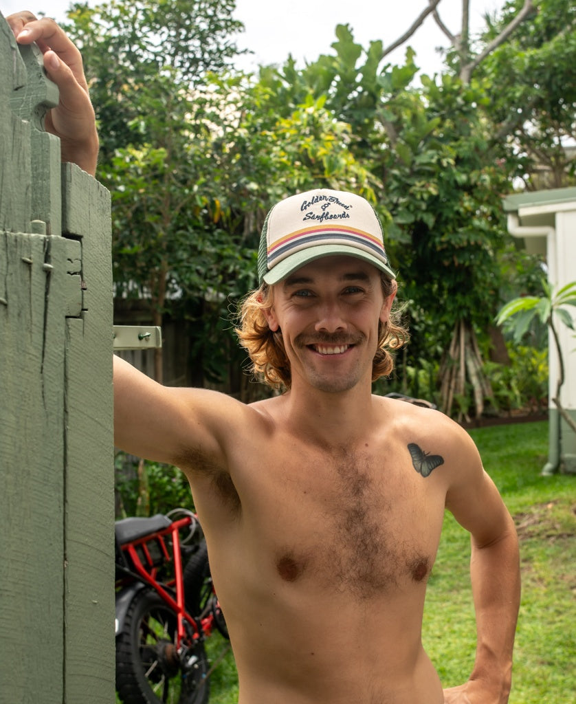 A mustached man in a Golden Breed GoJo Trucker Cap (Khaki) stands by a green fence, smiling. He has a butterfly tattoo on his chest; trees and a red bicycle are in the background.