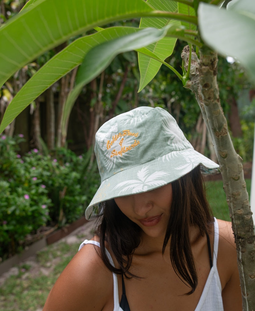 A woman stands outdoors near a tree, wearing the Golden Breed Chelsea Bucket Hat in Sage. The lightweight cotton linen fabric keeps her comfortable as she enjoys the fresh air surrounded by greenery.