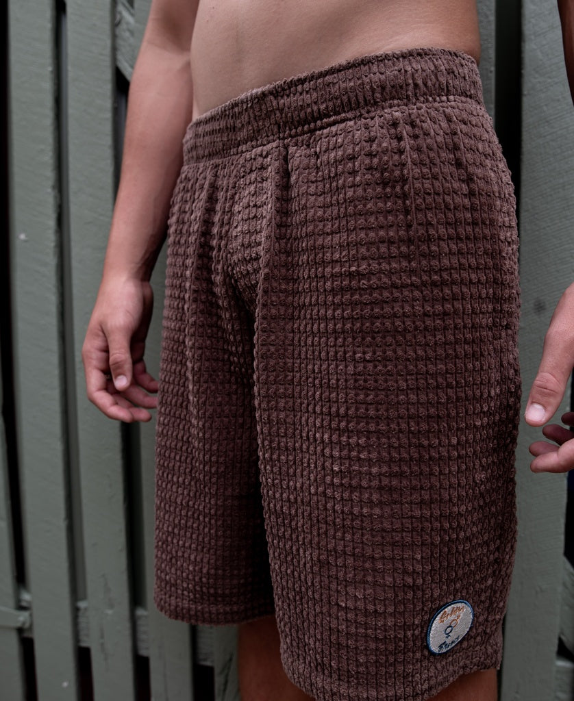 Someone wearing Golden Breeds El Solito Walkshort in Wash Brown, featuring a textured cotton cord and an elastic waist with a small patch on the lower left leg, stands before a green wooden fence.