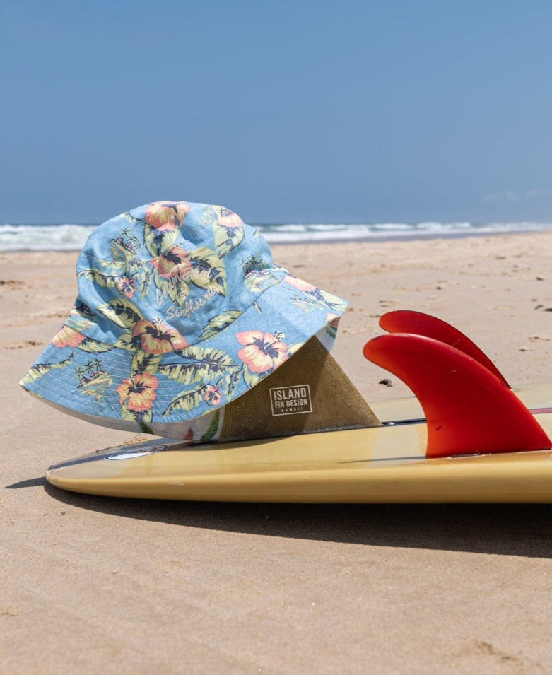 The Chelsea Bucket Hat in Azure Blue by Golden Breed sits on the fin of a yellow surfboard with red fins, resting on the sandy beach near the ocean.