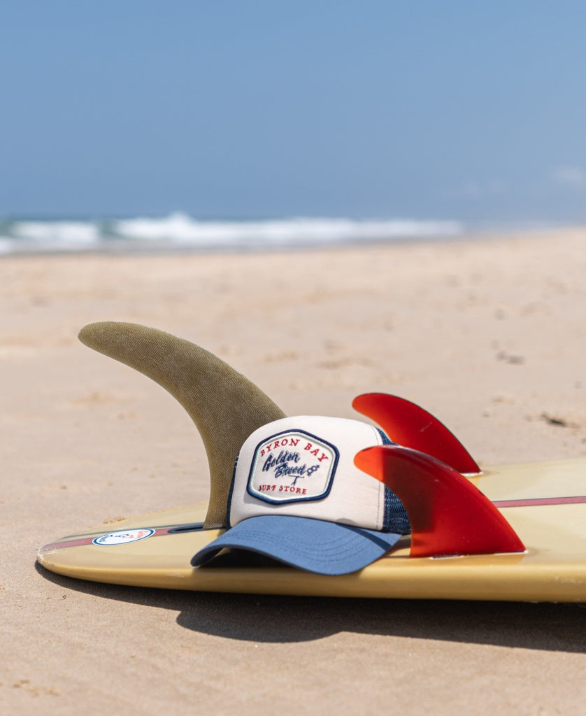 A Byron Trucker Cap | Dark Denim by Golden Breed rests on a surfboard with three fins on the sandy beach, as gentle ocean waves roll in the background.