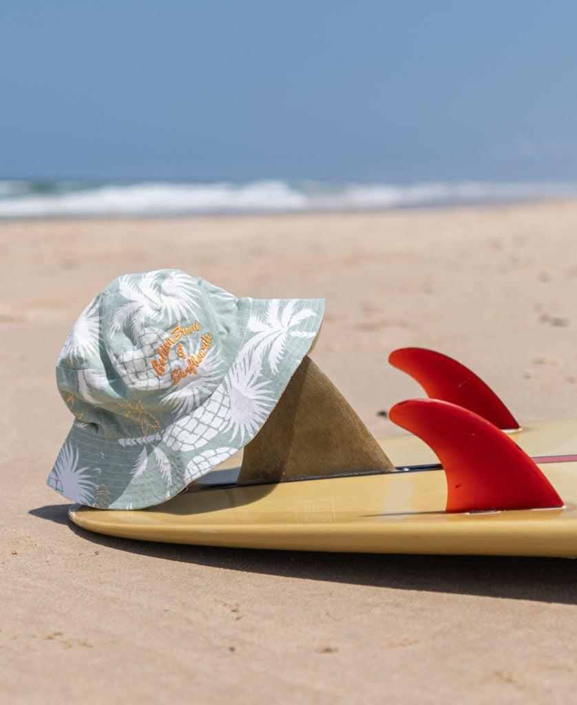 The Chelsea Bucket Hat | Sage by Golden Breed, featuring a light green shade with white palm prints, sits on a yellow surfboard with red fins on the sandy beach near the ocean.