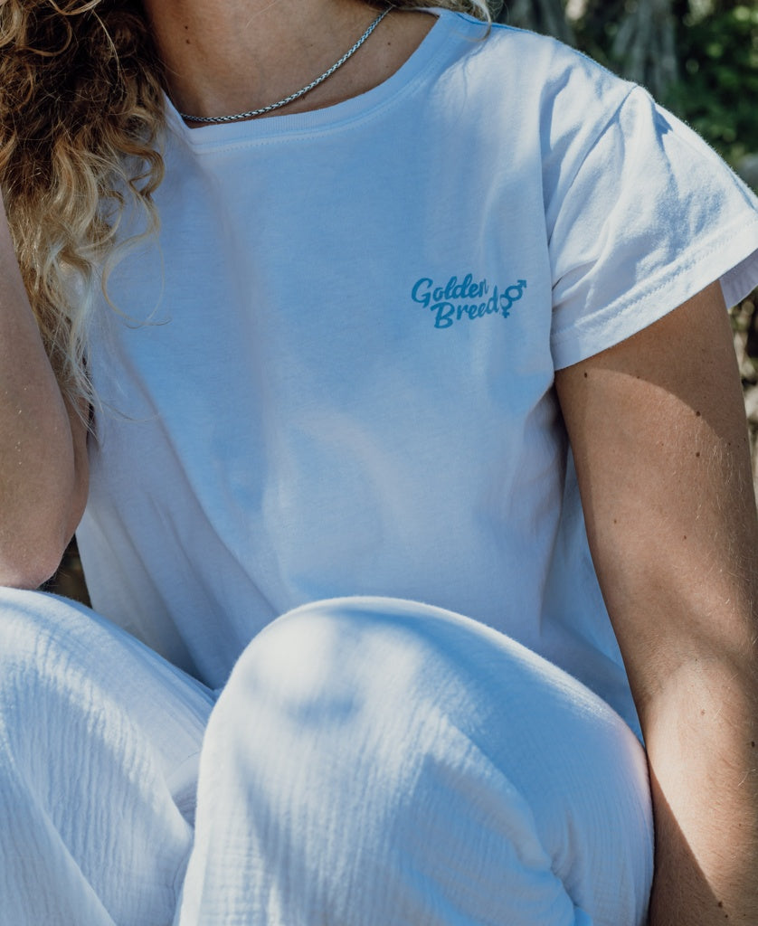 Wearing the Golden Breed Hattie Crop Tee in white and matching white pants, this person relaxes outdoors as sunlight filters through the trees in the background.
