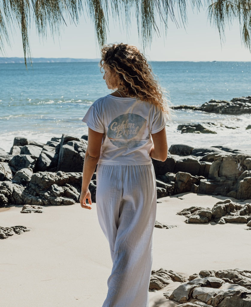 A person with curly hair stands on a rocky beach facing the ocean, wearing the Golden Breed Hattie Crop Tee in white paired with white pants.