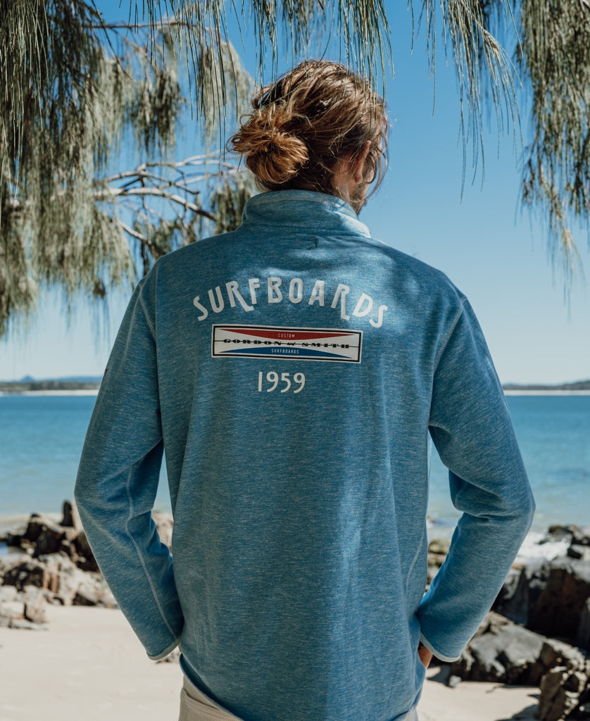 A person with long hair in a blue Gordon & Smith G&S 1/4 Zip Surfboards Rag Top | Sky Marle stands on a rocky beach, facing the ocean. Surfboards 1959 is printed on the back of the jacket.
