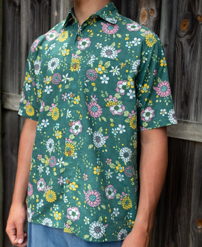 A person stands in front of a wooden fence wearing the Golden Breed Happy Floral | Ivy Green shirt, featuring a relaxed fit and vibrant floral pattern.