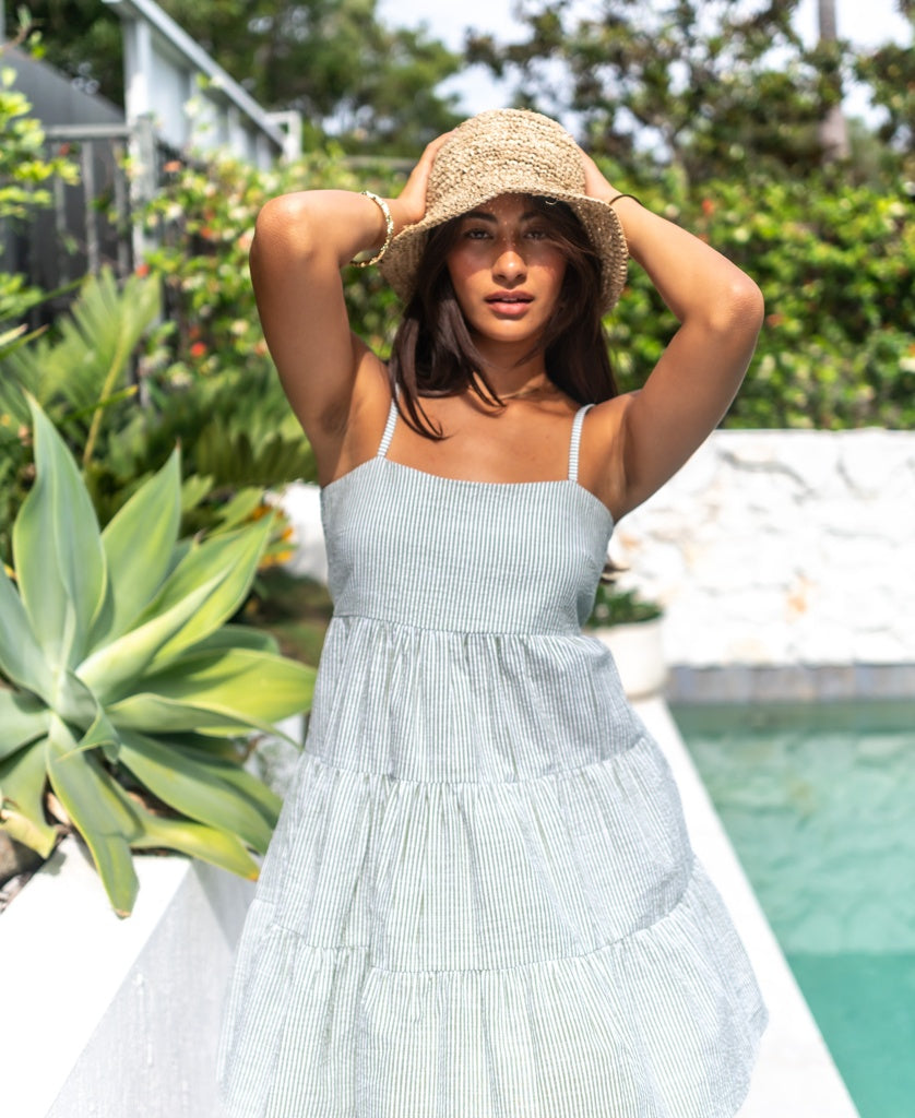 A woman in a light summer dress wears the Lazy Beach Raffia Bucket Hat | Natural by a swimming pool, holding her hat with both hands. Lush greenery surrounds her, capturing the essence of summer.