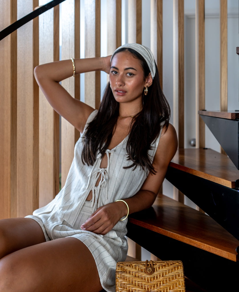 A woman sits on wooden stairs, wearing a light sleeveless outfit, gold jewelry, and a Lazy Beach Head Scarf | Fine Stripe handmade in Bali. A wicker handbag is beside her and vertical wooden slats form the background.
