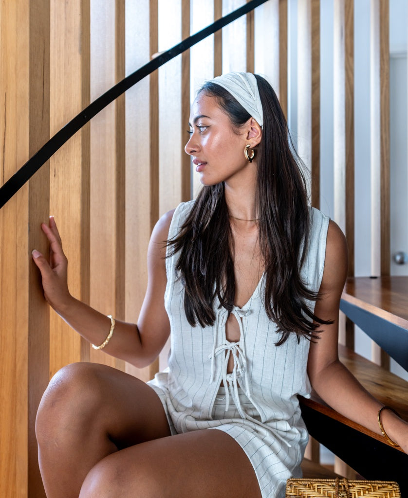 A woman with long dark hair wears the Lazy Beach Head Scarf | Fine Stripe, sitting on wooden stairs and looking to the side, her sleeveless striped outfit and handmade Bali accessory completing a stylish, summery look.