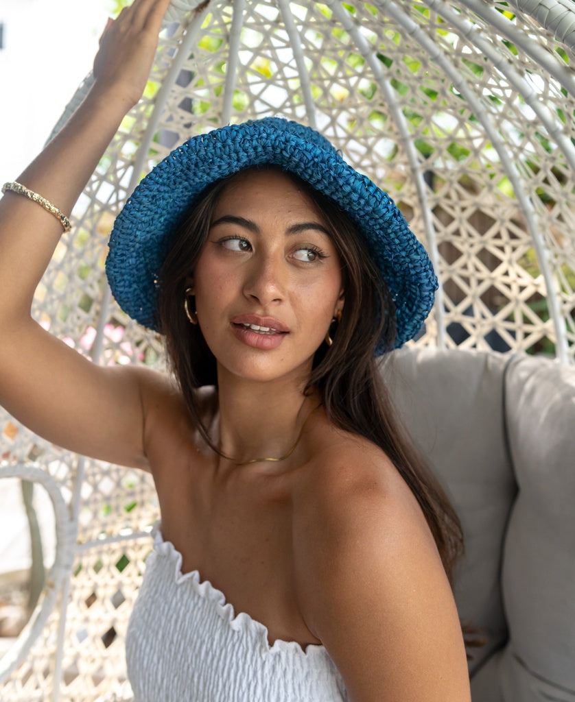 A woman in the Lazy Beach Raffia Bucket Hat with Brim in Blue sits on a wicker chair, wearing a white strapless top and looking to the side with a relaxed expression.