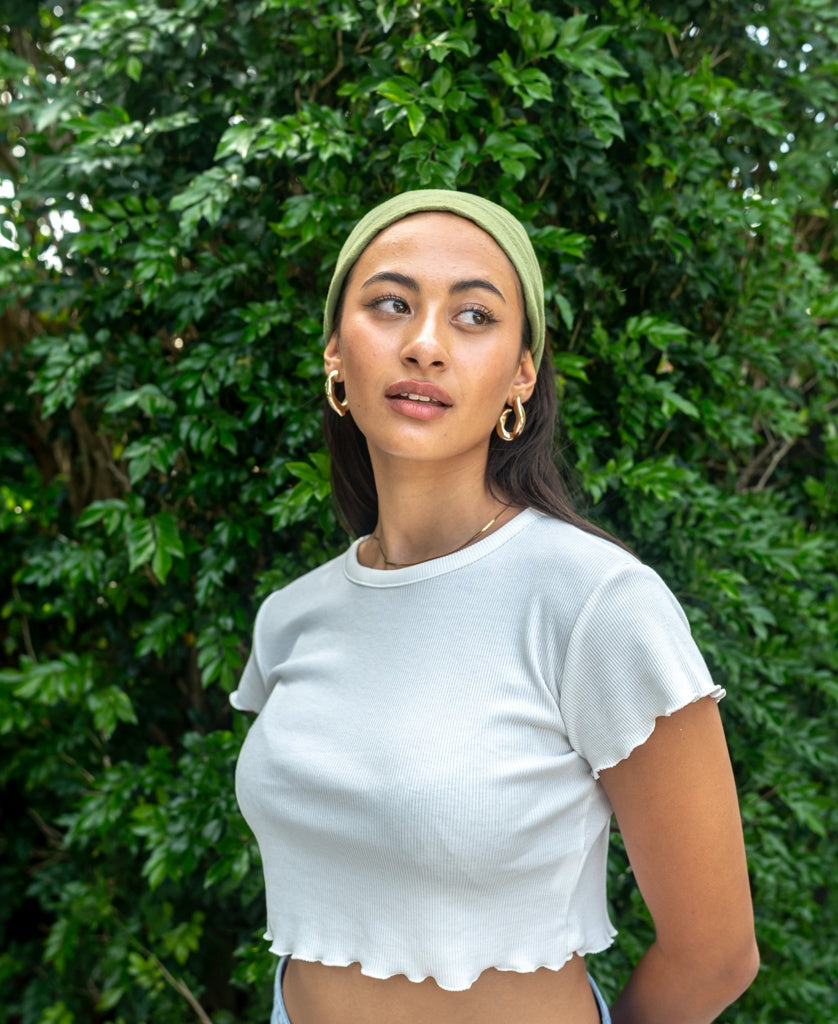 A woman wearing the Lazy Beach Head Scarf in Khaki and a white cropped shirt stands in front of green leafy bushes, looking off to the side.