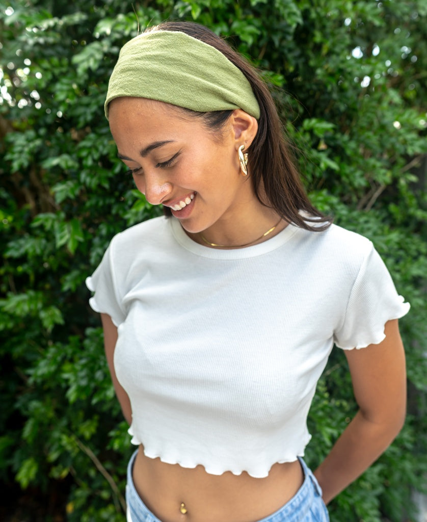A woman stands outdoors before green foliage, smiling and looking down while wearing a white cropped top, gold jewelry, and the Lazy Beach Head Scarf in Khaki—handmade in Bali.