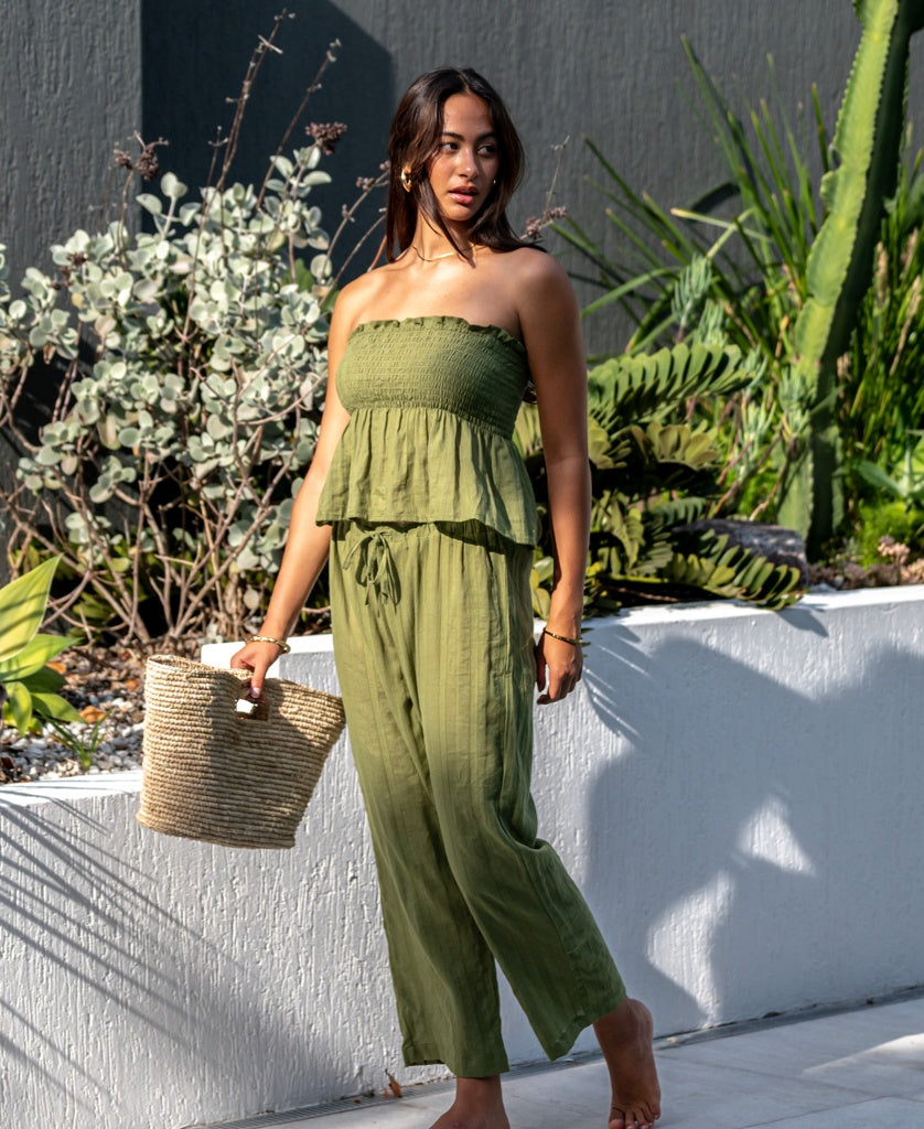 A woman stands outdoors barefoot, wearing the Lazy Beach Liberty Pant in Khaki with a matching green strapless top, holding a woven basket amid lush greenery.