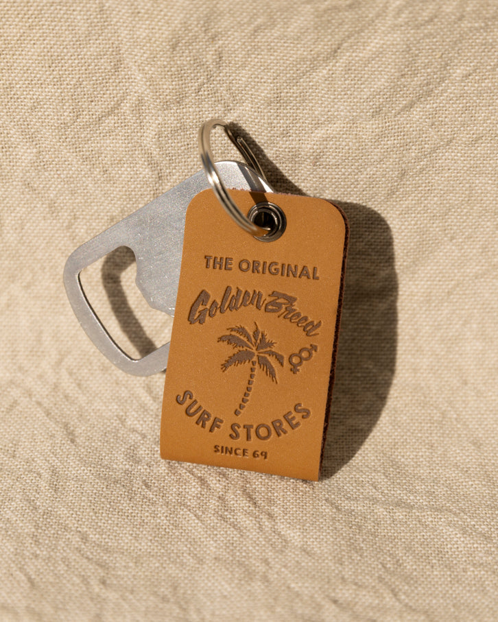 The Golden Breed GB Keyring combines a metal bottle opener and a brown leather keychain embossed with The Original Golden Breed Surf Stores Since 69 and a palm tree, adding classic surf style to your essentials.