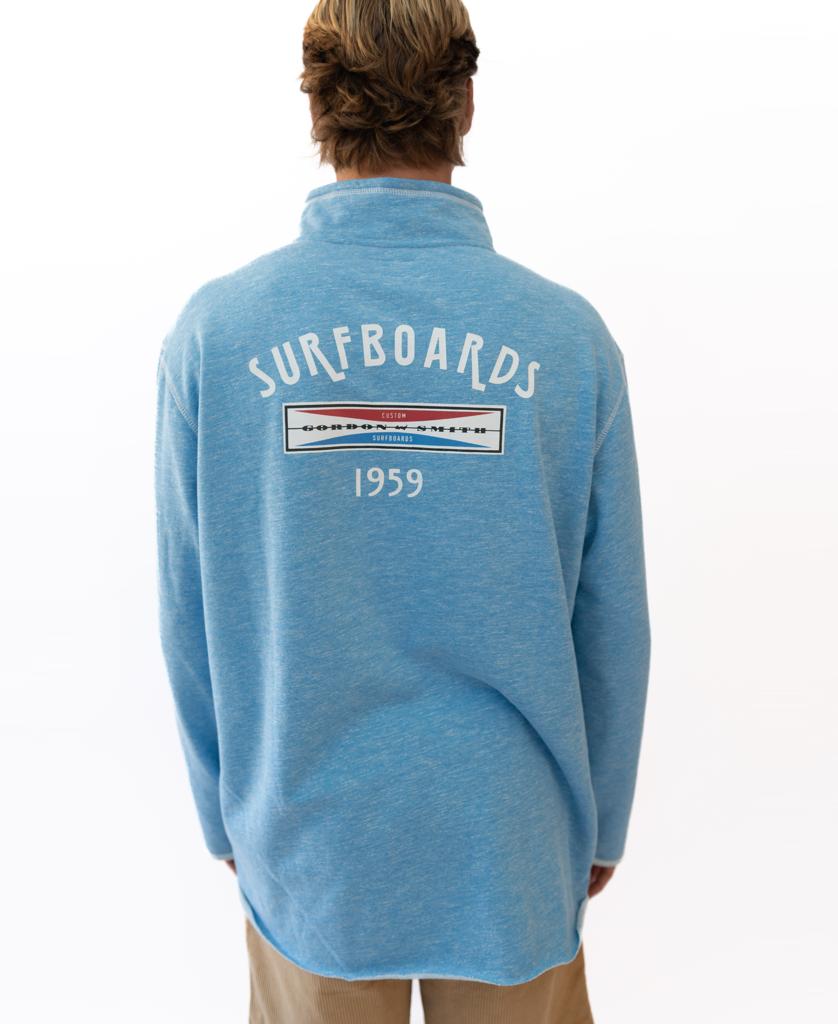 A person wears the Gordon & Smith G&S 1/4 Zip Surfboards Rag Top in Sky Marle, featuring SURFBOARDS 1959 and a rectangular logo on the back, standing against a plain white background.
