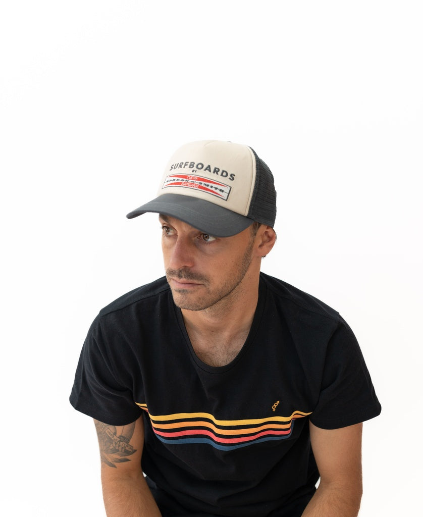 A man sits against a plain white background wearing a black t-shirt with colorful stripes and a Gordon & Smith G&S Boards Trucker Cap in wash black, featuring beige and black cotton canvas with logo embroidery.