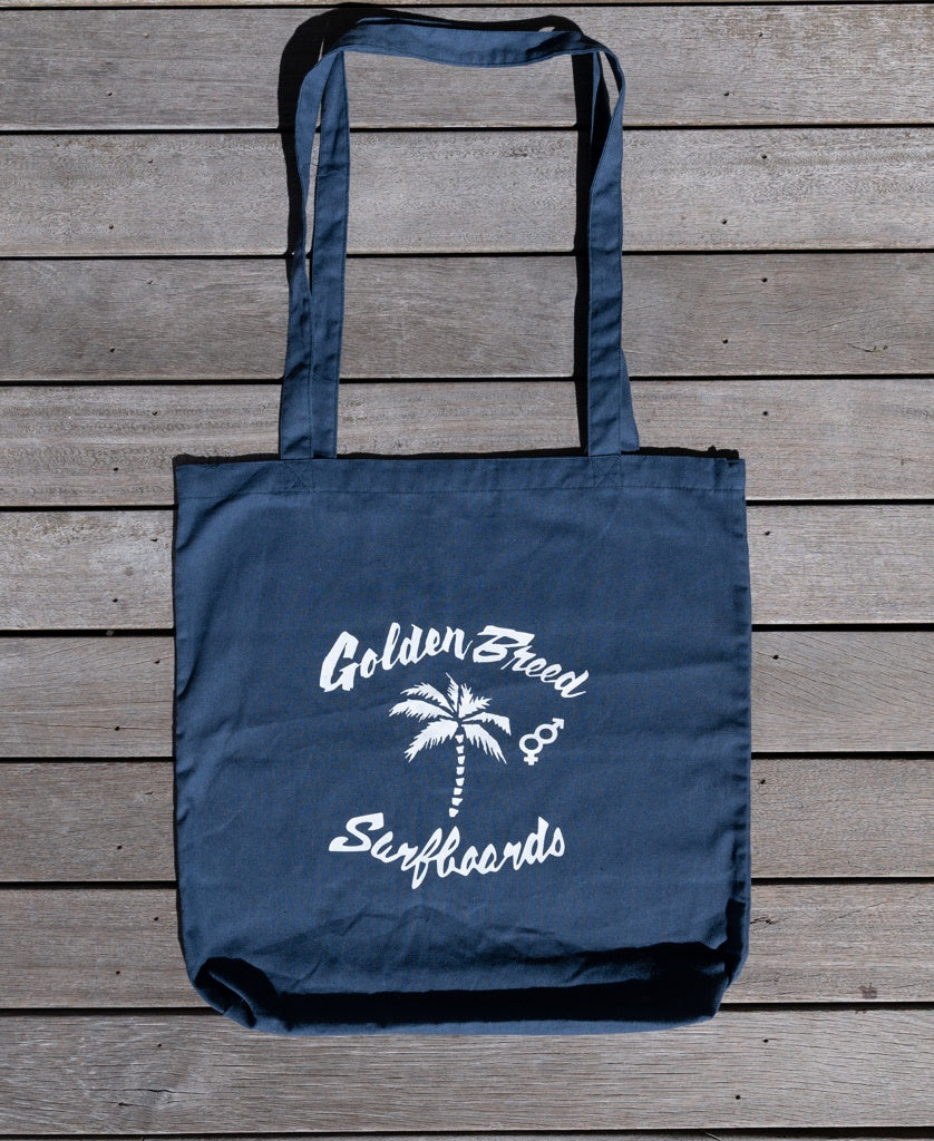 The Bardot Logo Tote Bag in vintage navy by Golden Breed features a white palm tree and surfboards graphic on the front and is displayed on a wooden surface.