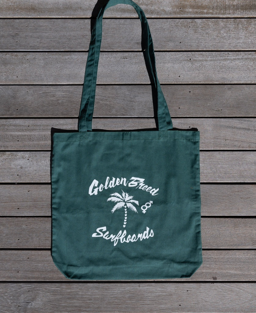 The Bardot Logo Tote Bag in Ivy Green by Golden Breed features white Golden Breed Surfboards text, a palm tree illustration, and logo details, displayed on wooden planks.
