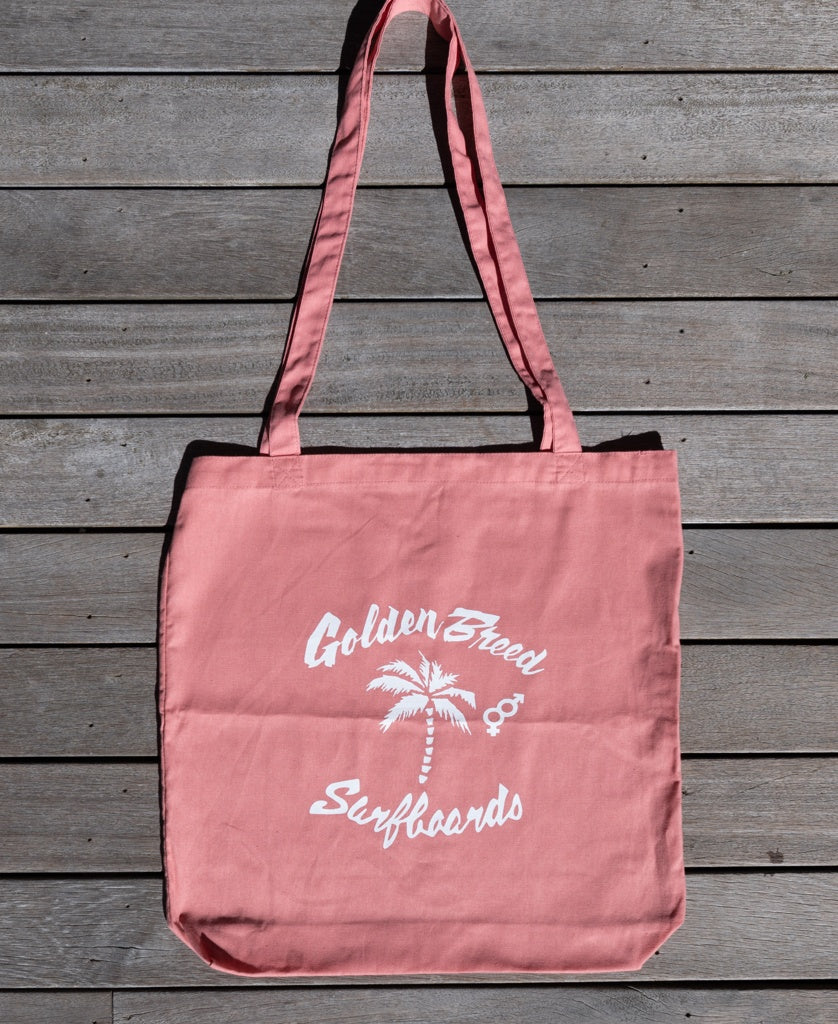The Bardot Logo Tote Bag | Rose by Golden Breed features a white palm tree and surfboards graphic on pink cotton. Ideal as a reusable shopping bag for everyday use. Shown here on a wooden surface.