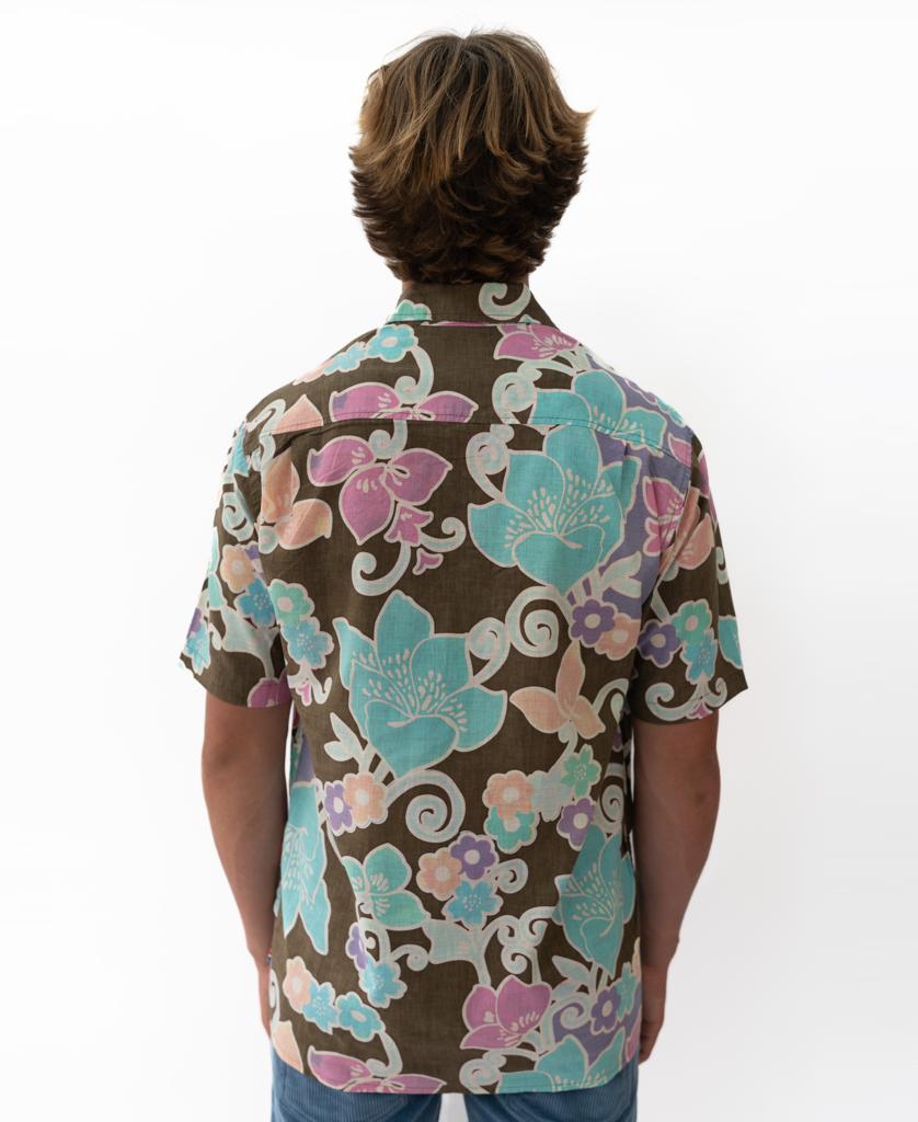 A person with medium-length hair faces away, wearing the Golden Breed Batik Shirt | Wash Brown, a short-sleeved shirt with a colorful floral pattern on a brown background.