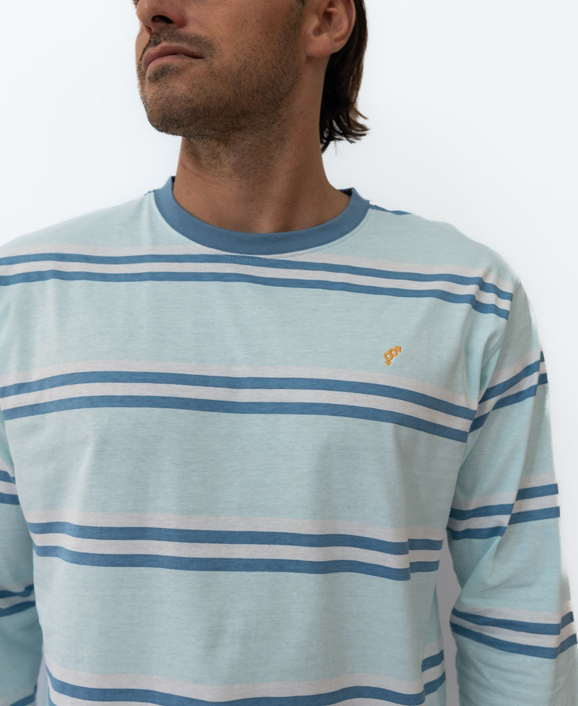 A man in the Golden Breed Bay Stripe LS Tee | Ice—a light blue, long-sleeve shirt with blue and white stripes—stands against a plain white background, capturing a relaxed beach surf vibe.
