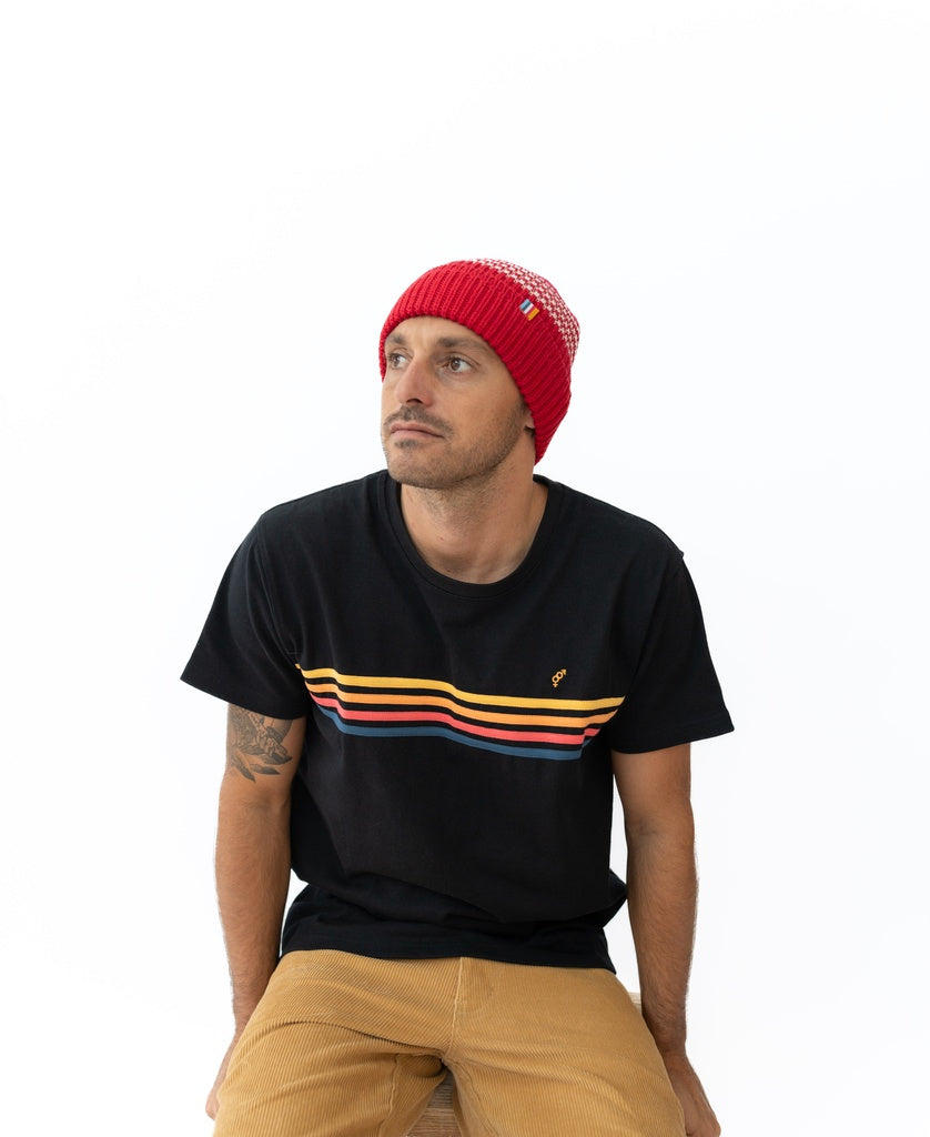 A man in the Golden Breed Beach Boy Wool Beanie in red, a black tee with colorful horizontal stripes, and tan pants sits on a stool against a plain white background.