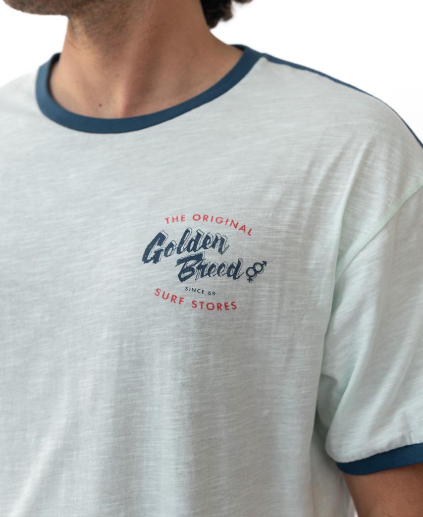 Someone wears the Golden Breed Break Ringer Tee | Ice, a light T-shirt with blue trim and a chest logo reading The Original Golden Breed Surf Stores Since 69, designed in Australia for classic comfort.