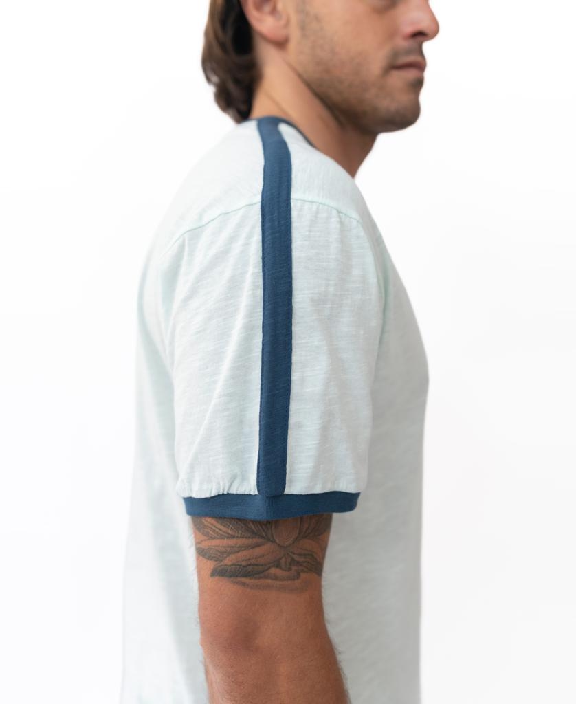 A man in profile wears the Golden Breed Break Ringer Tee | Ice, made from 100% cotton and designed in Australia, featuring dark blue trim on the sleeves and shoulder while showcasing a floral tattoo on his upper arm.