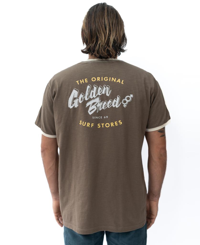 A person with medium-length hair wears a brown Golden Breed Break Ringer Tee featuring white and yellow Golden Breed Surf Stores text printed on the back.