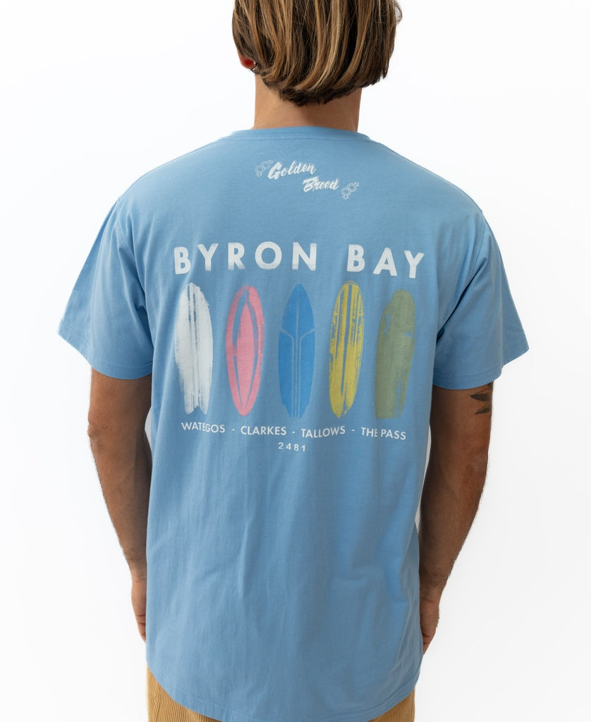 A person wearing the Golden Breed Byron Boards Tee in Azure Blue, crafted from soft cotton jersey, featuring surfboard graphics and BYRON BAY with four iconic Surf Town beaches listed on the back.