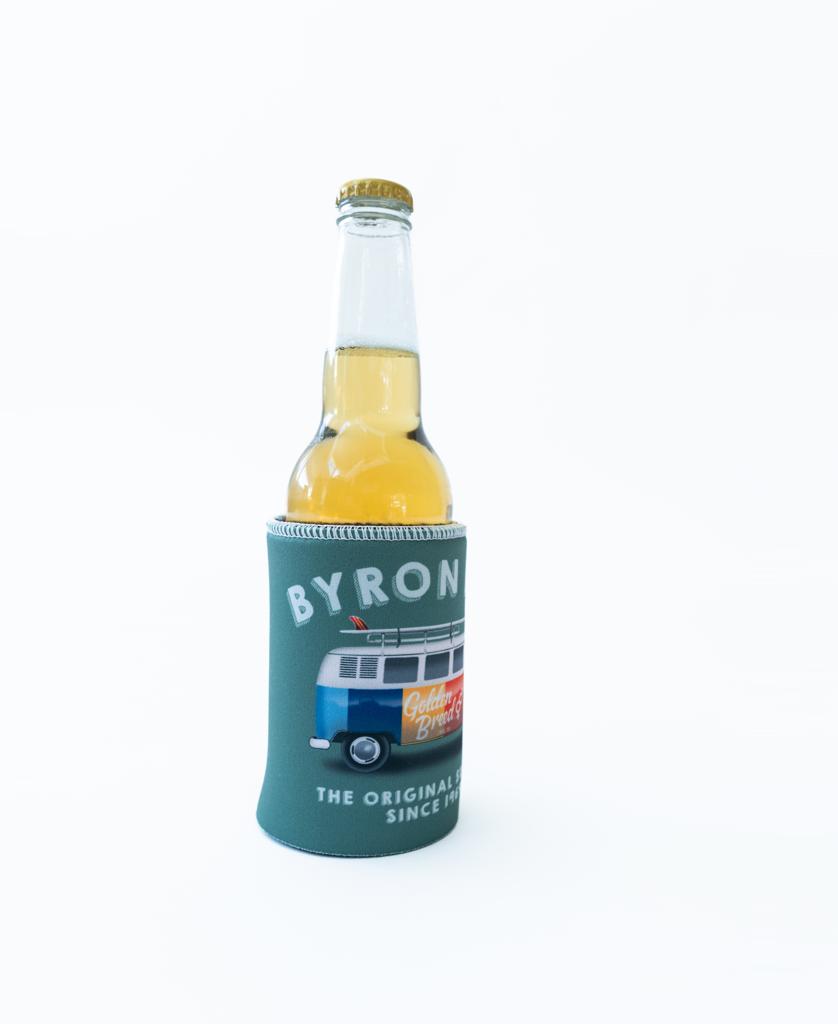 A light beverage in a glass bottle sits in the Golden Breed Byron Stubby Kombi | Surf Green holder, featuring a van design and BYRON BAY text, on a plain white background.