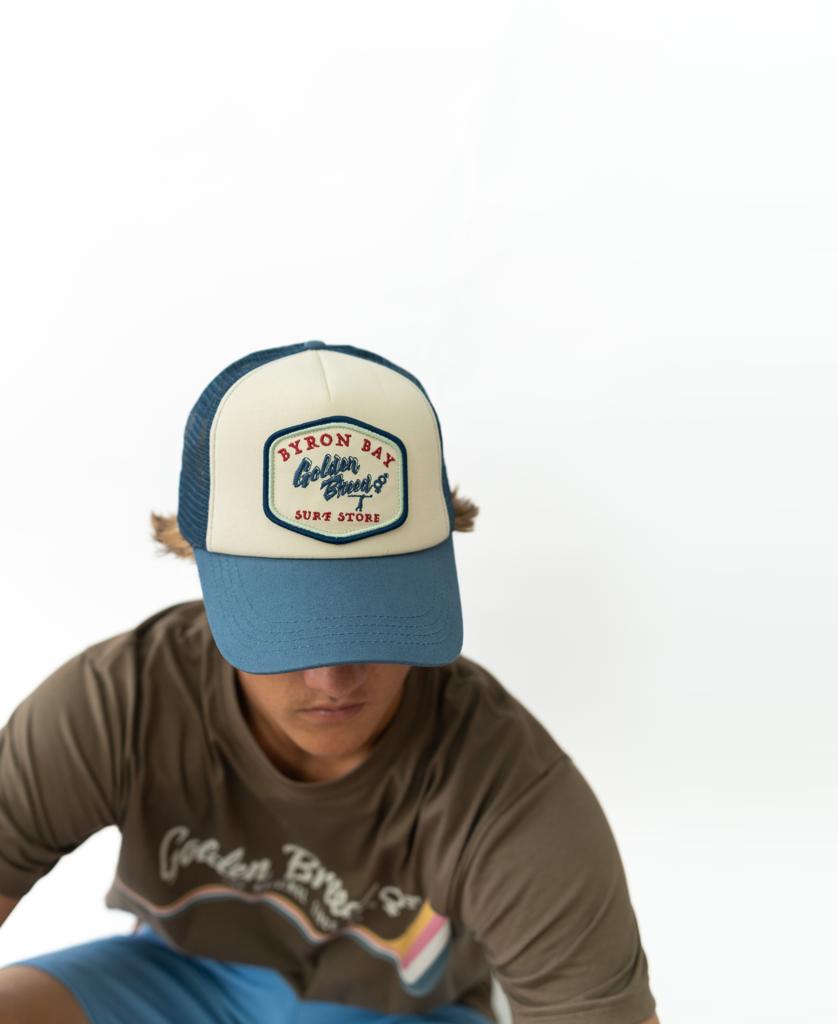 A unisex look featuring Golden Breeds Byron Trucker Cap in Dark Denim, paired with a brown graphic tee and dark denim jeans, seated against a plain white background.