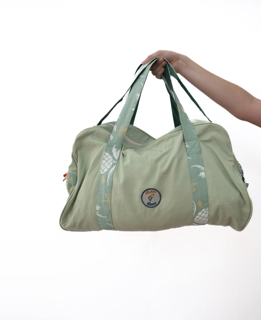 A hand holds the Golden Breed Pine Palm Canvas Roll Bag in Sage, featuring patterned straps and a circular patch on quality heavy cotton canvas, set against a white background.