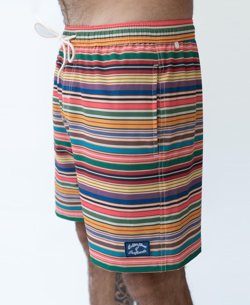 A person wears Golden Breed’s Daze Elastic Waist Boardie | Stripe—multicolored striped shorts with 4-way stretch, a white drawstring, and a small rectangular patch above the hem.