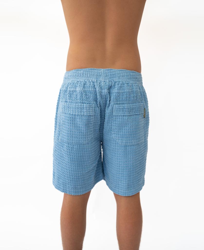 A person is seen from behind wearing Golden Breeds El Solito Walkshort in Azure Blue, crafted from cotton cord, with a plain white background.